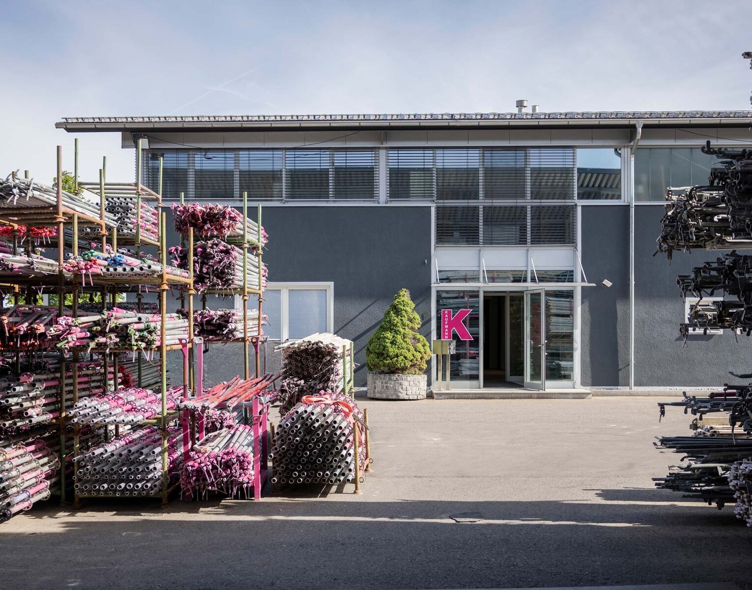 Gerüstbau Kaufmann Außenbereich eines Industriebetriebs mit gelagerten Metallrohren und einem Gebäude mit großem Eingang und einem rosa Buchstaben K an der Wand.
