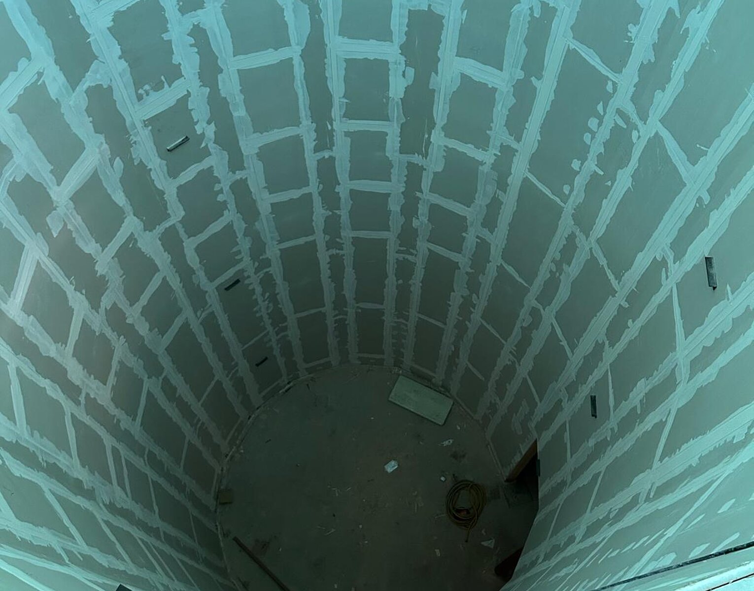 Blick nach unten in einen runden, unvollendeten Schacht mit Wänden aus Gipskartonplatten und sichtbaren Fugen, Boden mit Bauresten.