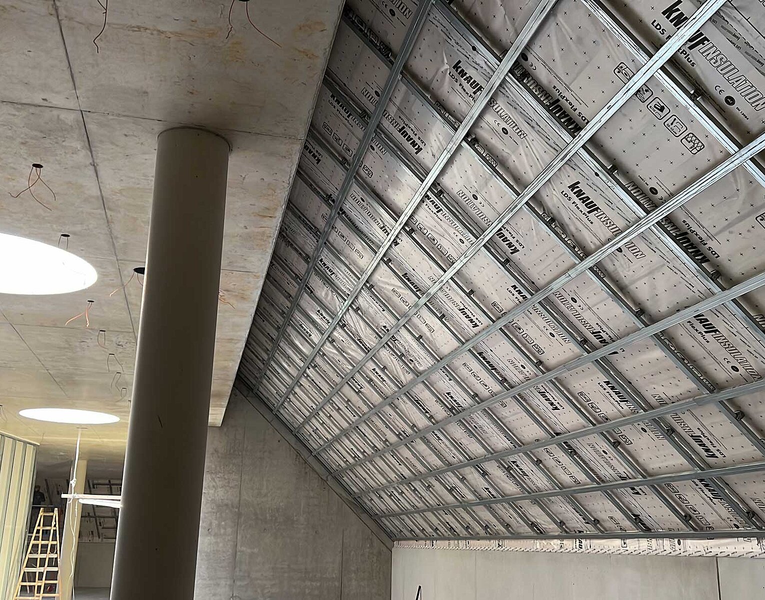 Blick in einen Raum mit Betondecke und einer schrägen Wand, an der eine Unterkonstruktion für Dämmplatten und Verkleidung installiert ist. Im Raum ist eine runde Säule und helle Deckenleuchten.