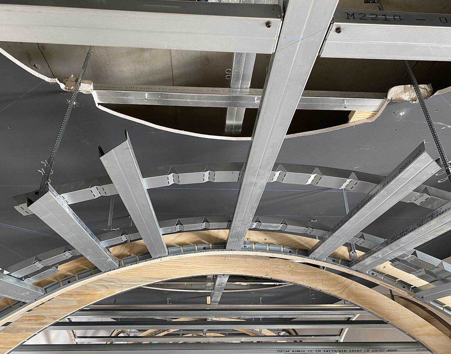 Innenaufnahme einer Deckenstruktur mit sichtbaren Metallträgern und hölzernen gebogenen Balken im Rohbaustadium.
