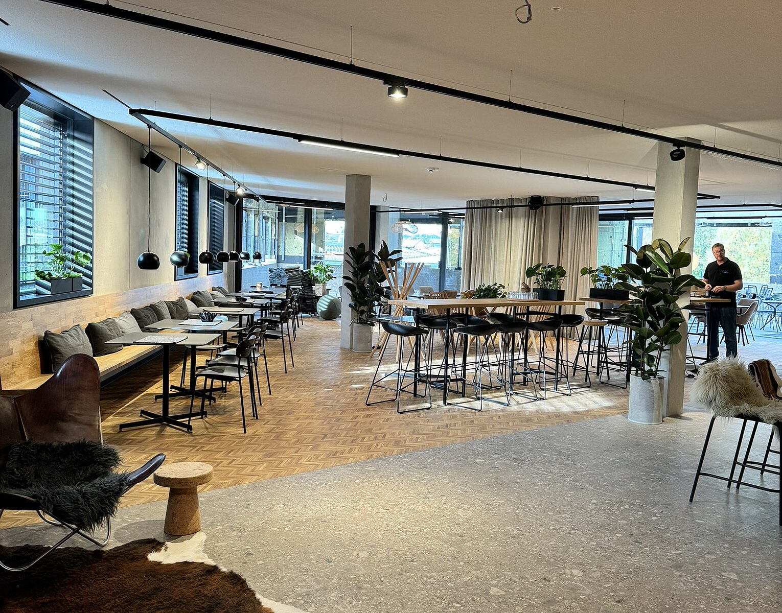Moderner, heller Coworking- oder Gemeinschaftsraum mit Sitzgelegenheiten am Fenster, hohen Tischen, Pflanzen und einem offenen Raumkonzept.