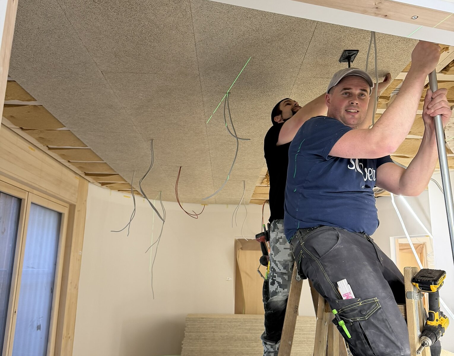 Zwei Handwerker montieren eine Akustikdecke in einem Raum, einer steht auf einer Leiter und hält eine Leiste, der andere arbeitet im Hintergrund an der Decke.