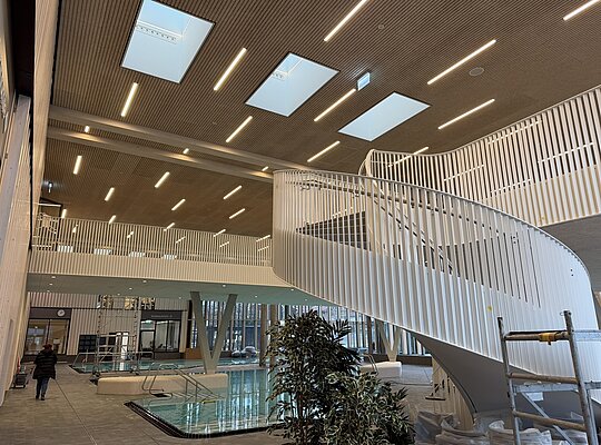 Trockenbau Kaufmann Therme Obersdorf Innenansicht eines modernen Schwimmbads mit einem geschwungenen weißen Treppengeländer, mehreren Wasserbecken, großen Fensterfronten und einem Holzdecken mit integrierten Deckenleuchten.