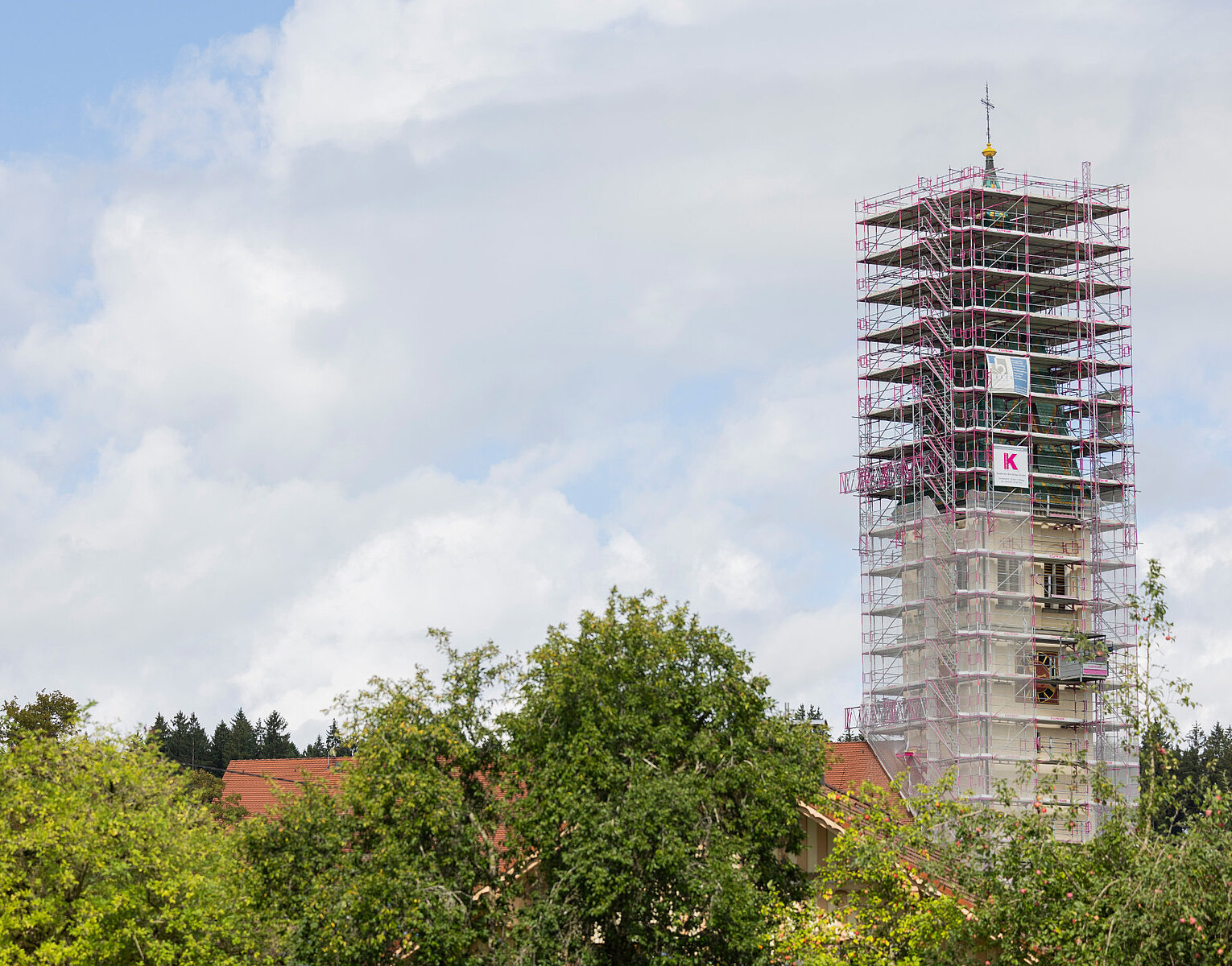 Ein hoher Turm, der von einem Gerüst umgeben ist, umgeben von Bäumen und unter einem bewölkten Himmel.