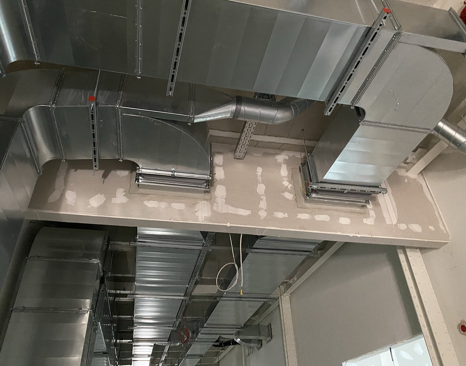 Innenaufnahme eines Gebäudes mit sichtbaren metallischen Lüftungskanälen und Rohren an der Decke und Wand in einem Rohbauzustand.