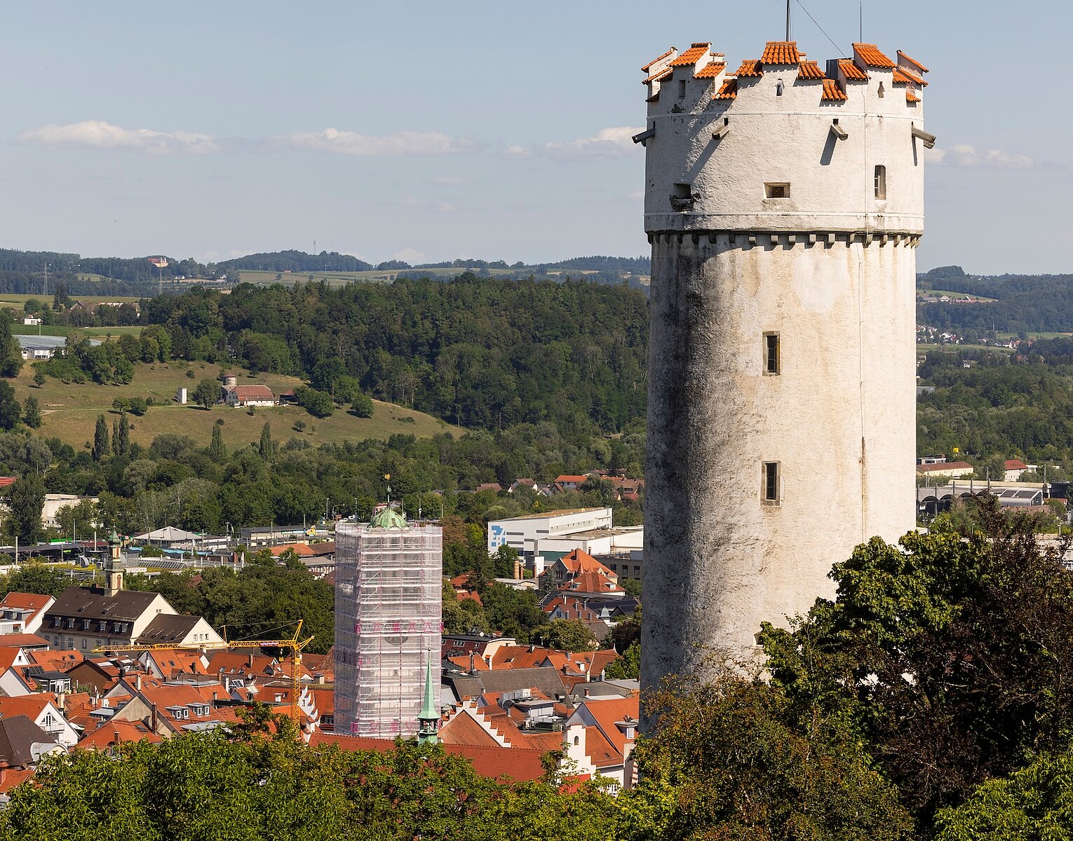 Weißer mittelalterlicher Turm mit Zinnen in einer Stadtlandschaft, umgeben von Häusern und grünen Hügeln im Hintergrund.