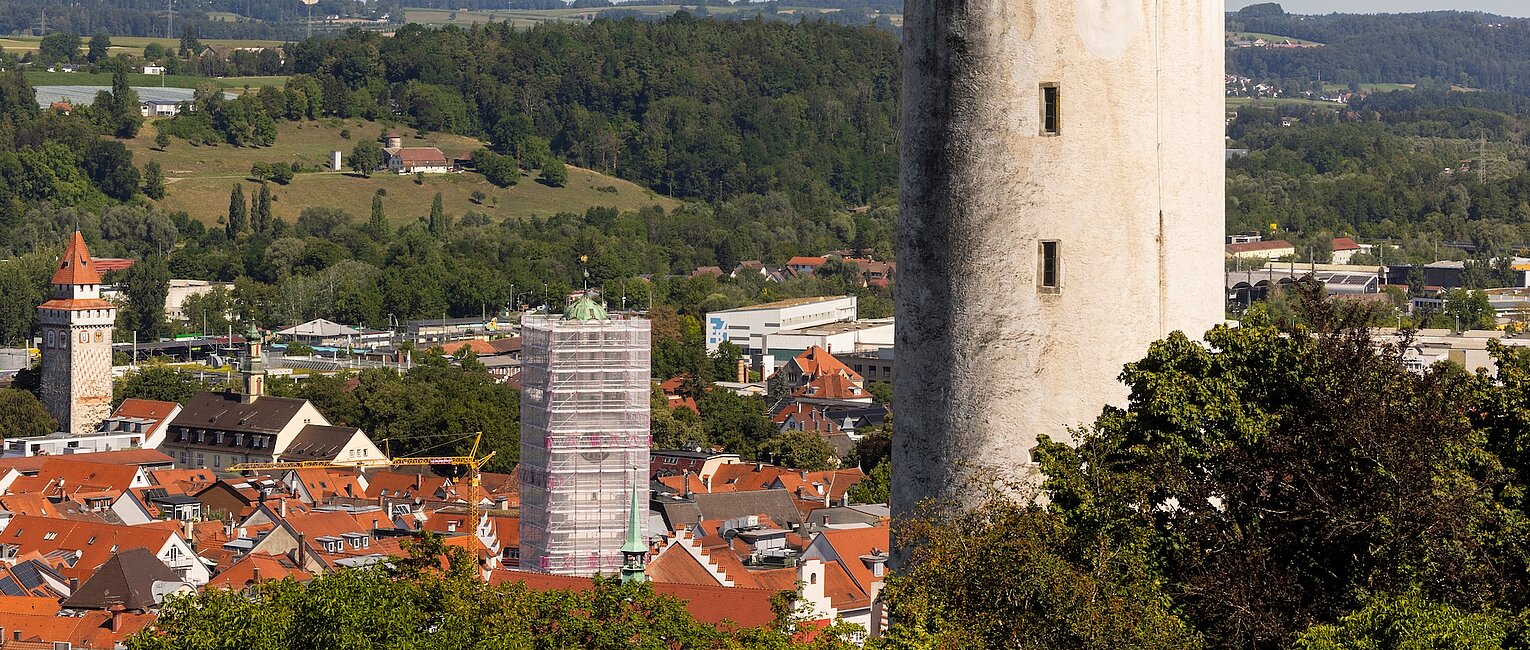 Weißer mittelalterlicher Turm mit Zinnen in einer Stadtlandschaft, umgeben von Häusern und grünen Hügeln im Hintergrund.