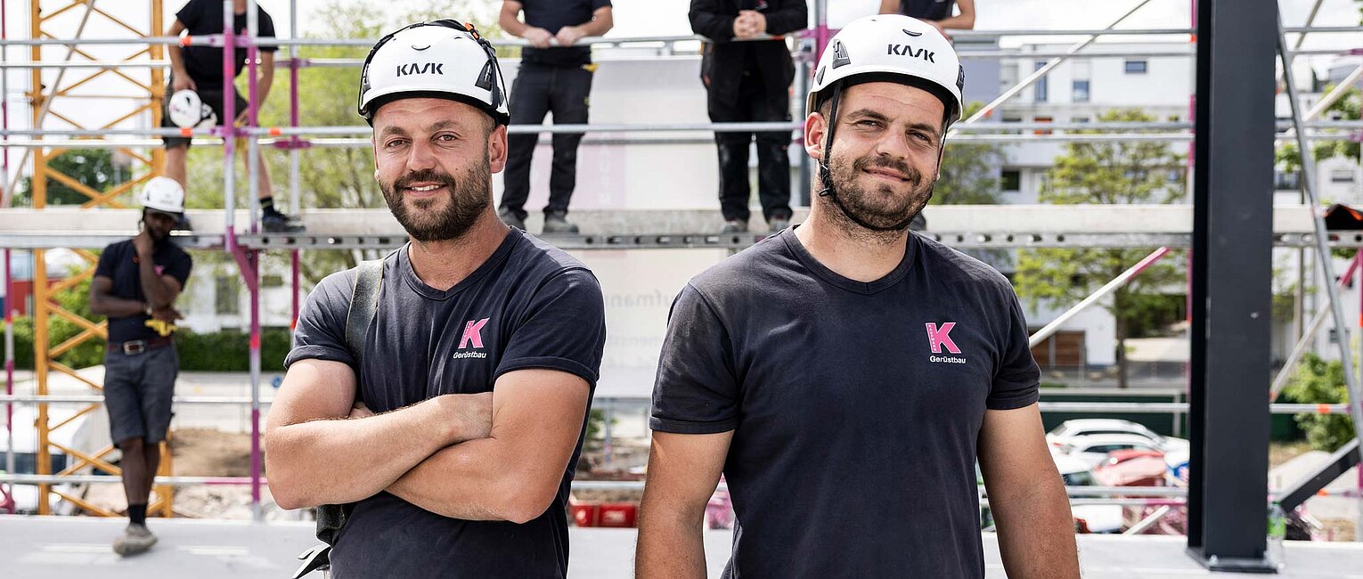 Zwei Bauarbeiter mit Schutzhelmen und schwarzer Arbeitskleidung stehen vor einer Baustelle mit Gerüst, weitere Bauarbeiter sind im Hintergrund sichtbar.