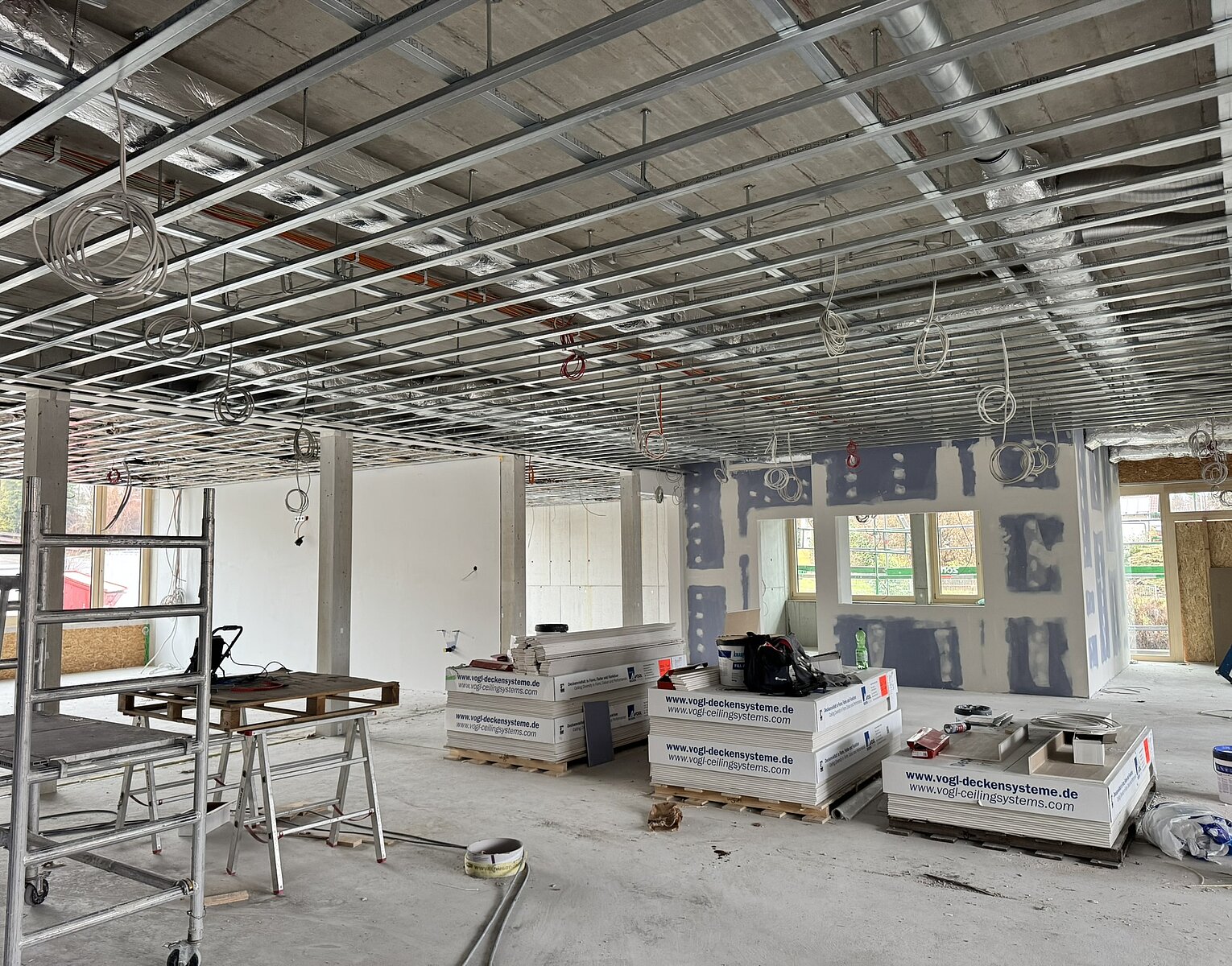 Innenraum einer Baustelle mit sichtbarer Deckenunterkonstruktion, gestapelten Gipskartonplatten und Baugerüst, teilweise verputzten Wänden und Baumaterialien auf dem Boden.