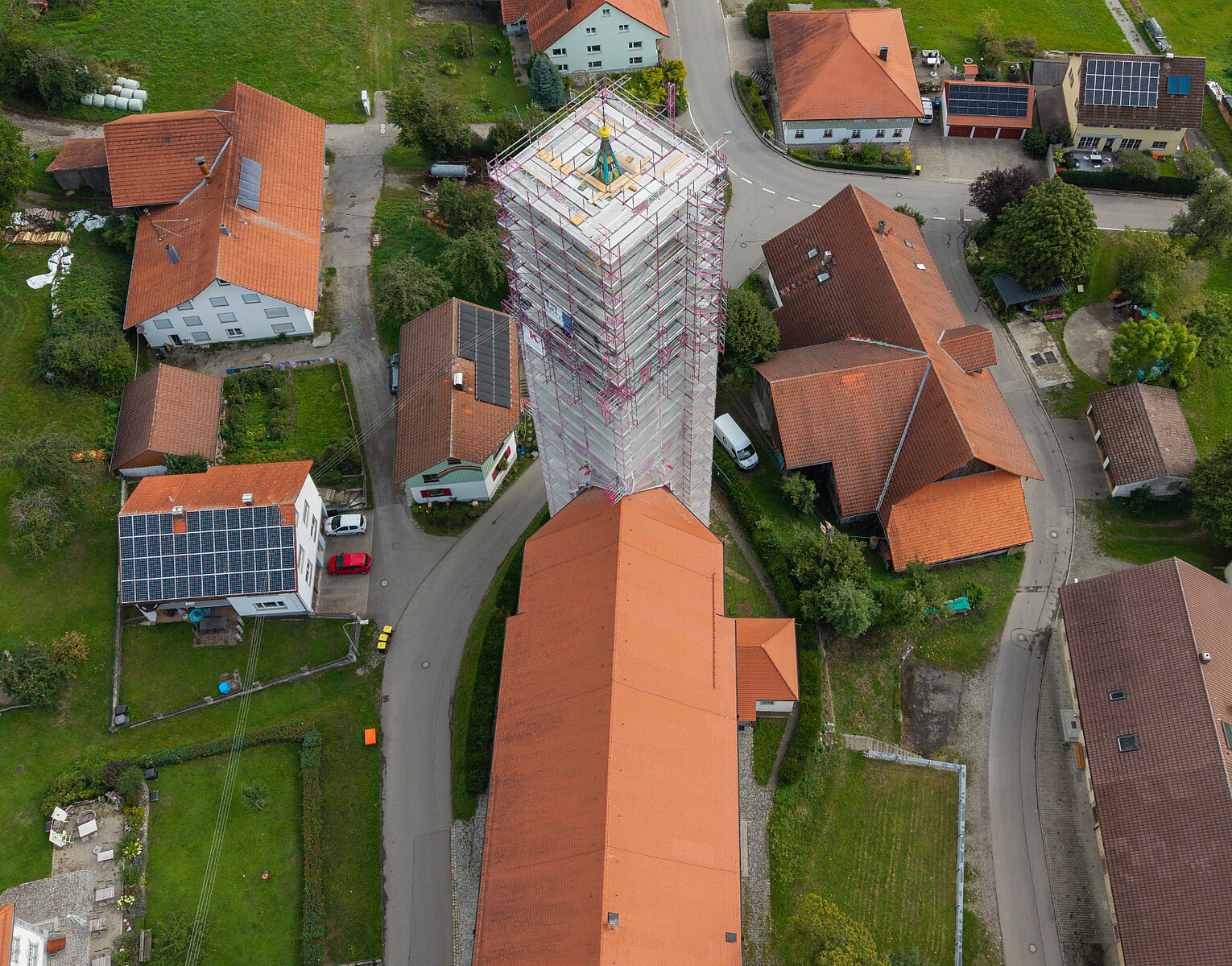 Luftaufnahme eines kleinen Dorfes mit Gebäuden und einem Turm in Gerüstbauweise, umgeben von Straßen und Grünflächen.