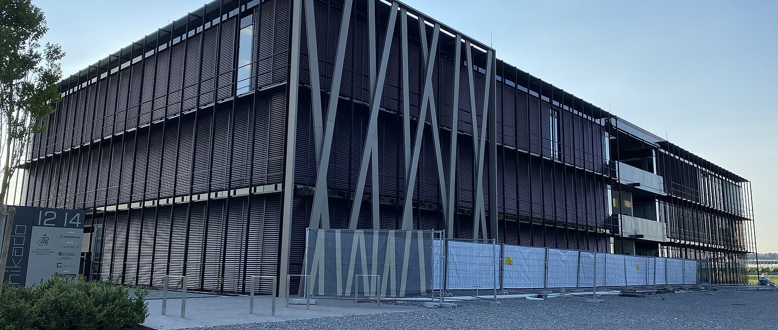Modernes, großes Bürogebäude mit dunkler Fassadenverkleidung und diagonal verlaufenden Stützen, umgeben von einem Kiesplatz und Bauzaun, bei klarem Himmel am Abend.