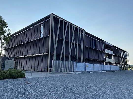 Trockenbau Kaufmann Mikado Friedrichshafen Modernes, großes Bürogebäude mit dunkler Fassadenverkleidung und diagonal verlaufenden Stützen, umgeben von einem Kiesplatz und Bauzaun, bei klarem Himmel am Abend.