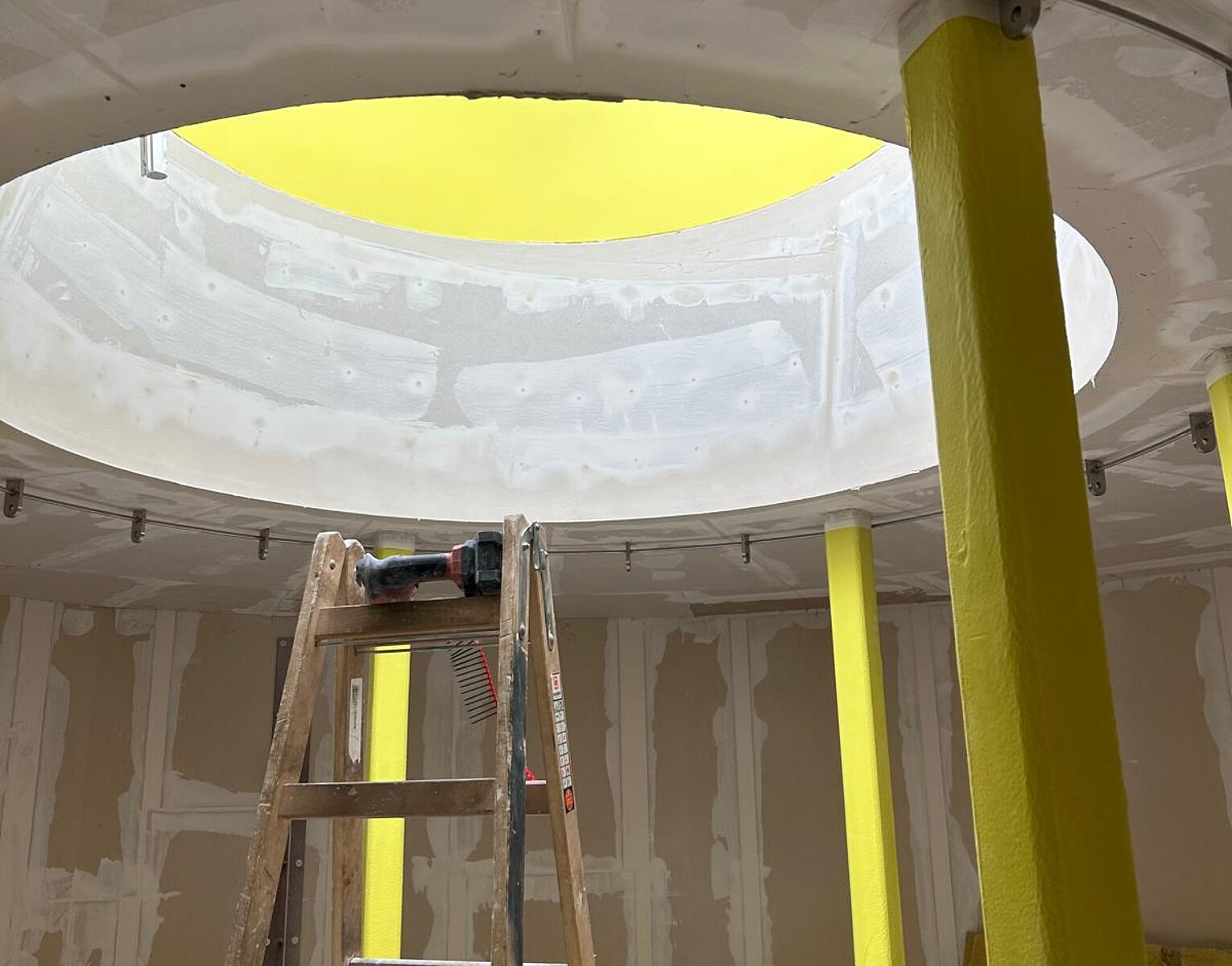 Innenraum einer Baustelle mit gelb gestrichenen Stützen, einer Leiter und einer runden Öffnung in der Decke, durch die natürliches Licht einfällt.