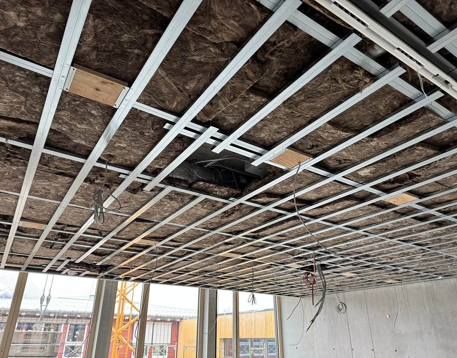 Decke eines Rohbaus mit Dämmung und Metallrahmen, Kabel hängen von der Decke, große Fenster und Baustelle im Hintergrund.