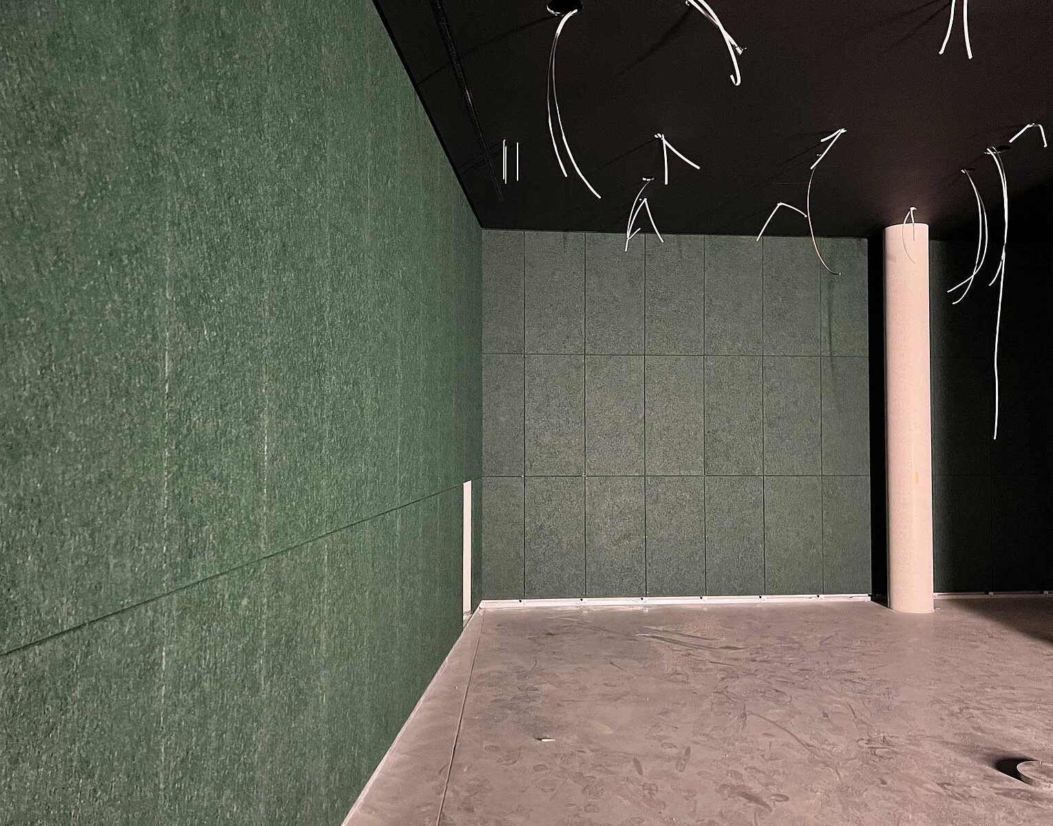 Leerstehender Raum mit grünen Wandpaneelen, grauem Boden und schwarzer Decke mit herabhängenden weißen Kabeln, in dem eine weiße Säule steht.