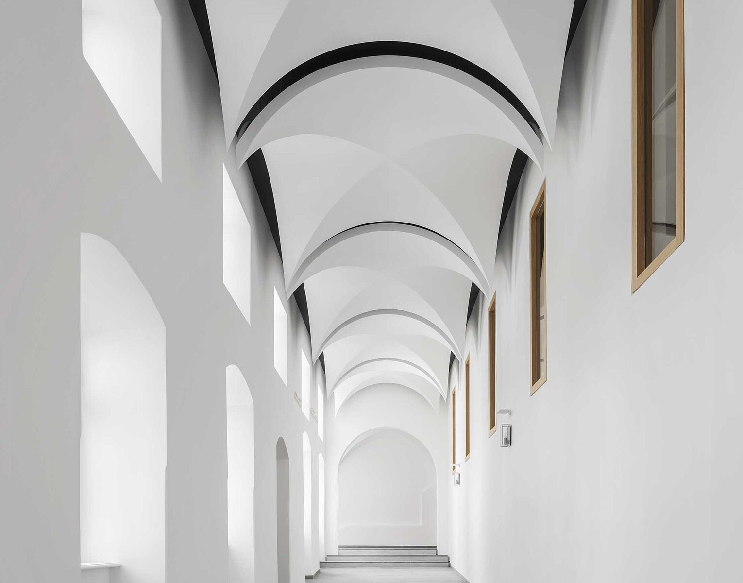 Blick in einen langen, weißen Gang mit gewölbter Decke und mehreren rechteckigen Fenstern an den Wänden auf beiden Seiten.