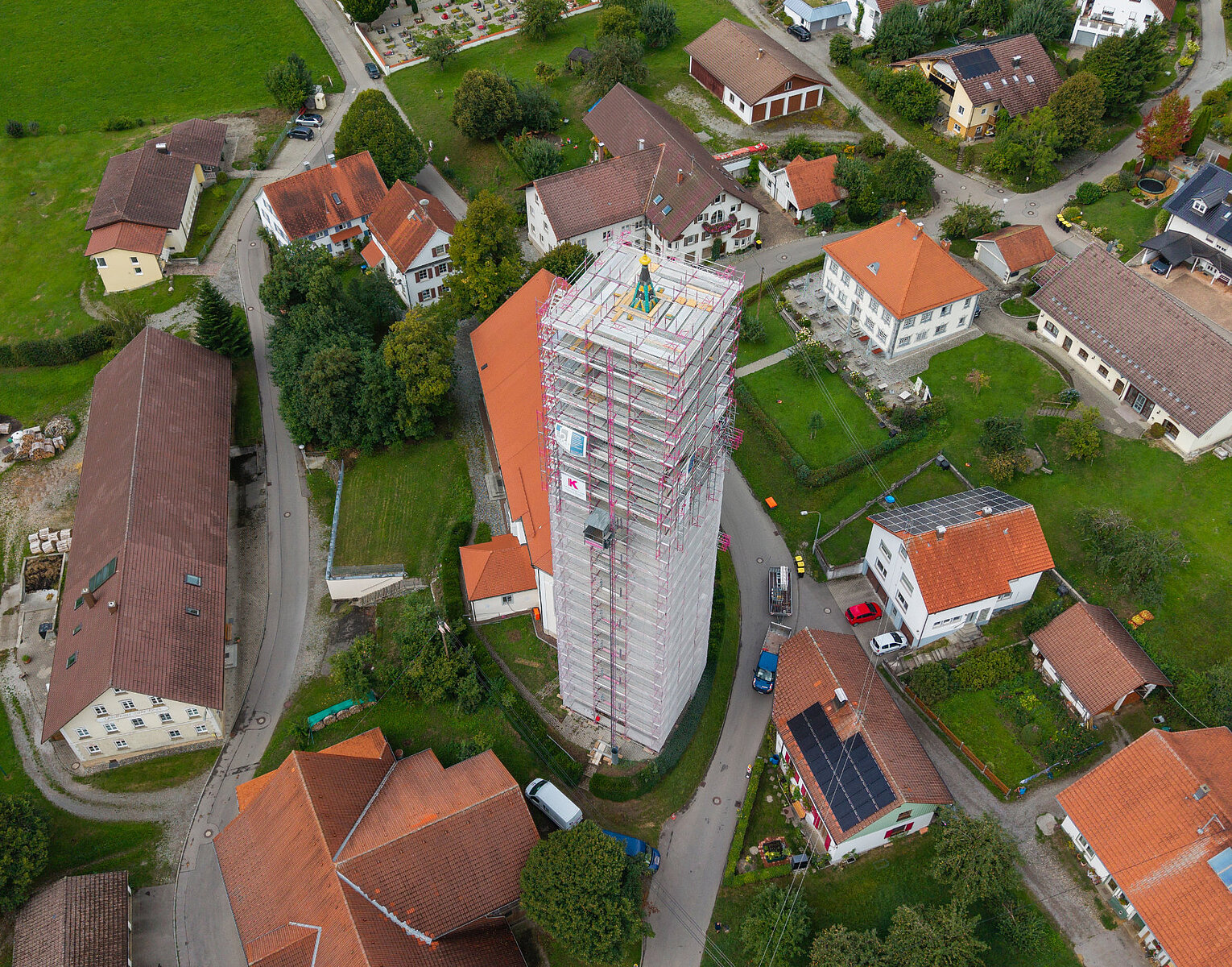 Luftaufnahme eines Turms mit Schutzgerüst während Renovierungsarbeiten, umgeben von mehreren Wohnhäusern in einer grünen ländlichen Umgebung.