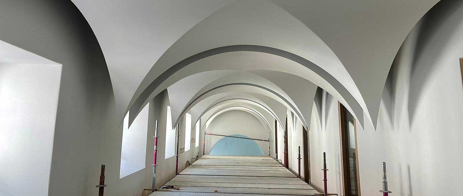 Langer Flur mit weißen, gewölbten Decken und großen Fenstern auf der linken Seite, Baugerüste entlang der Wände, helle Innenraumgestaltung.