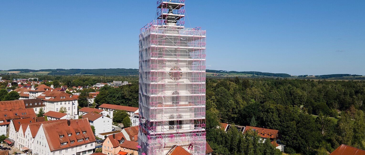Ein Kirchturm ist vollständig von einem Gerüst mit Netz umgeben, umgeben von Wohnhäusern und viel Grün unter einem klaren blauen Himmel.