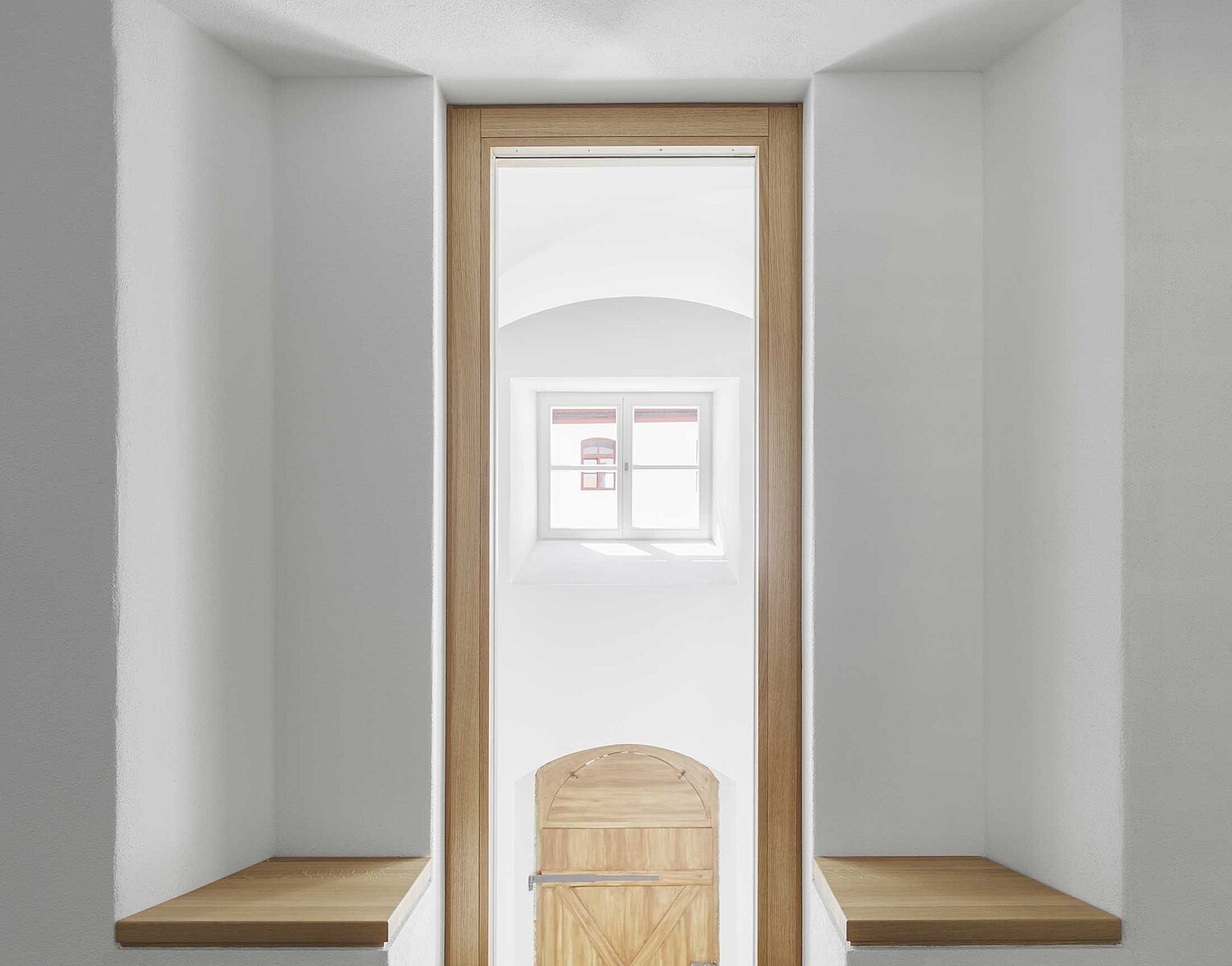 Minimalistisches helles Zimmer mit zwei integrierten, symmetrischen Holzbänken neben einem Fenster mit Blick auf eine Holztür und ein kleines Fenster dahinter.