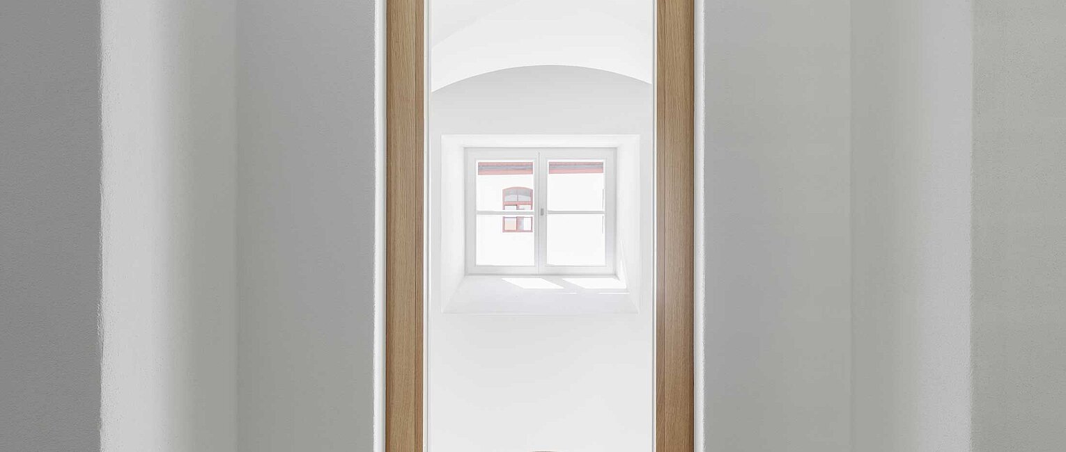 Minimalistisches helles Zimmer mit zwei integrierten, symmetrischen Holzbänken neben einem Fenster mit Blick auf eine Holztür und ein kleines Fenster dahinter.