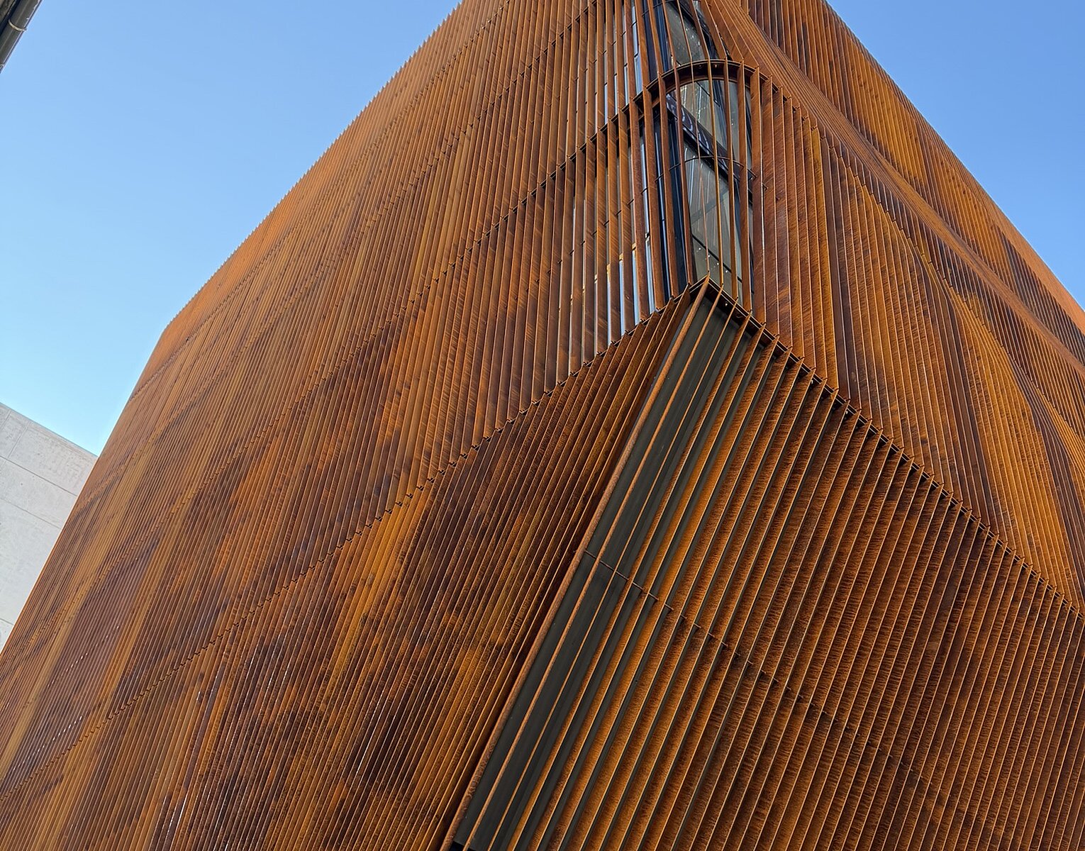 Blick auf eine moderne Gebäudeecke mit vertikalen, rostfarbenen Metalllamellen vor blauem Himmel.
