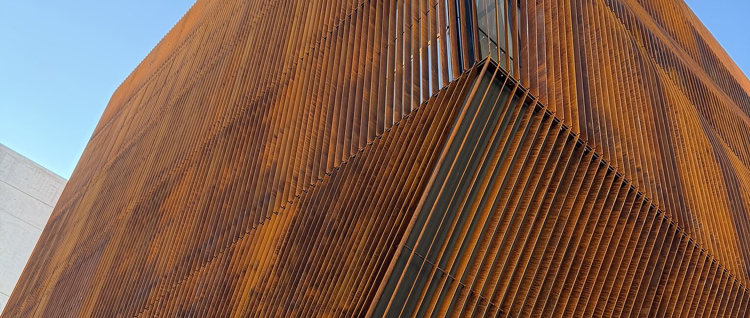 Blick auf eine moderne Gebäudeecke mit vertikalen, rostfarbenen Metalllamellen vor blauem Himmel.
