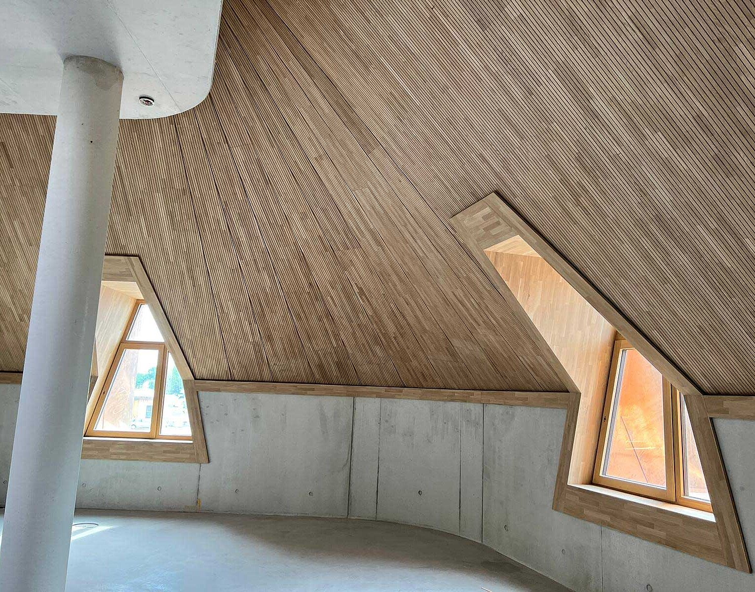Innenraum mit schrägen Holzdecken und zwei dreieckigen Fenstern mit Holzrahmen, stützender weißer Säule in der Mitte, Wände aus Beton, Tageslicht fällt ein.
