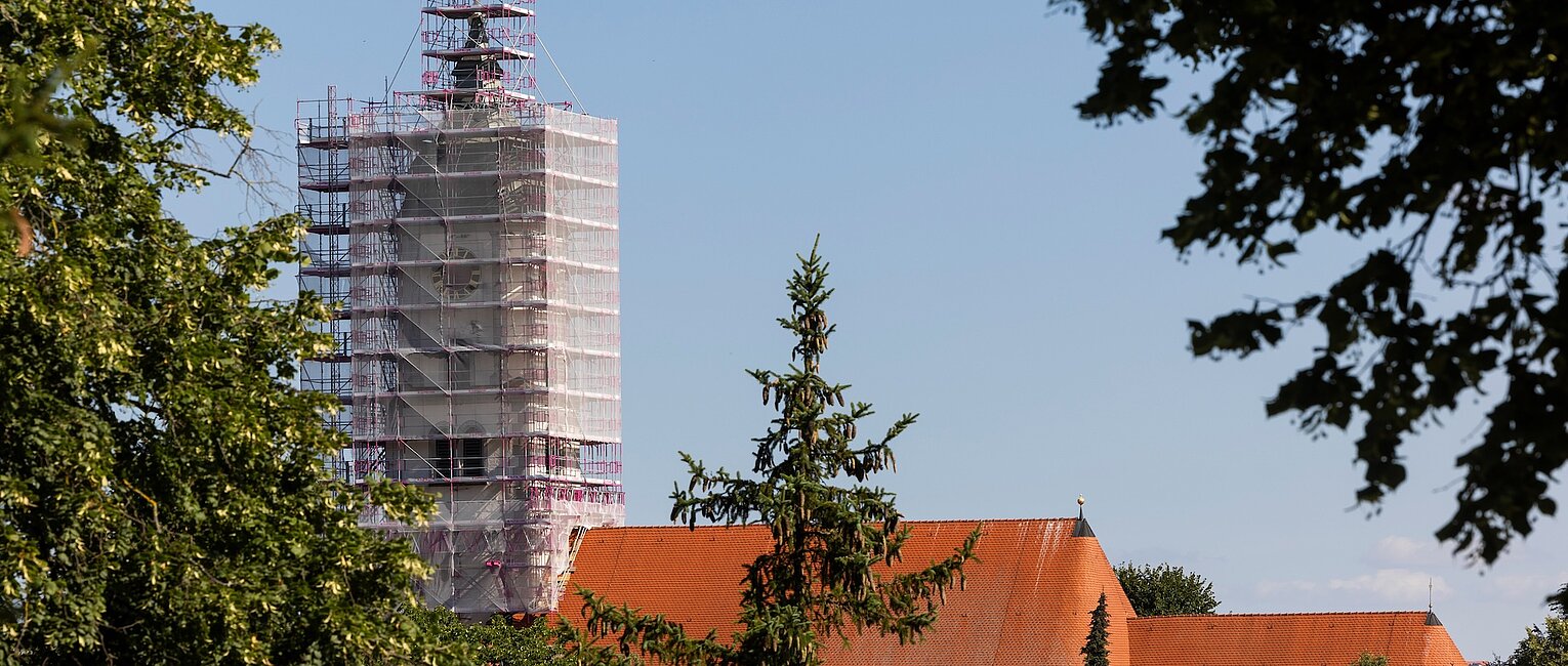 Turm eines Gebäudes, eingehüllt in ein Gerüst, vor klarem blauem Himmel, umgeben von grünen Bäumen und roten Dächern.