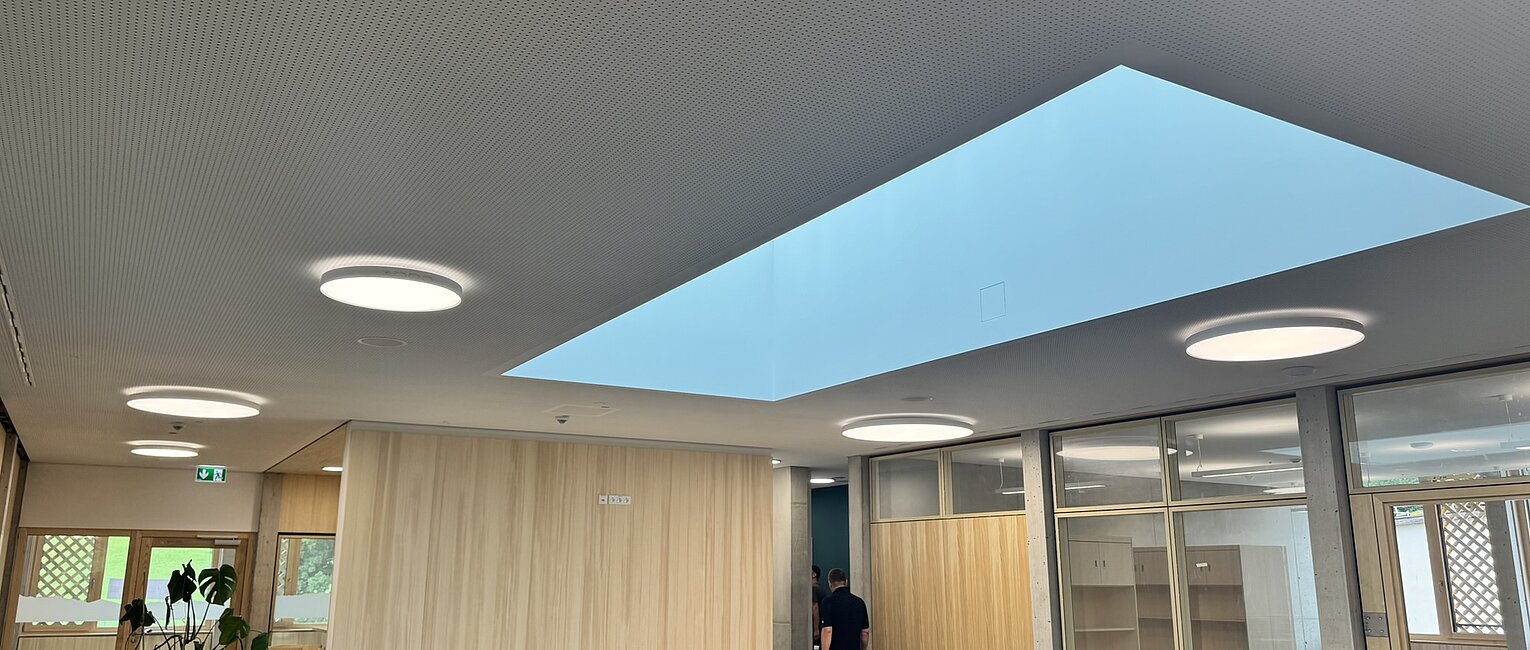 Büro mit Holzverkleidung, großem rechteckigem Oberlicht und runden Deckenleuchten, es sind Möbel und Personen im Hintergrund sichtbar.