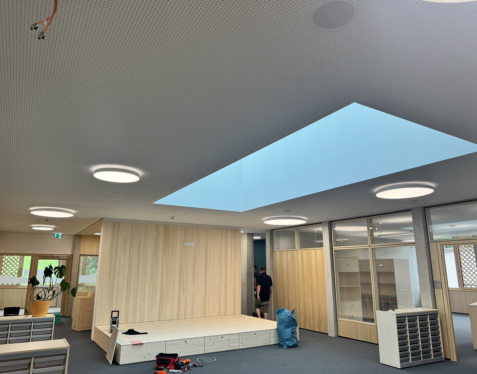 Büro mit Holzverkleidung, großem rechteckigem Oberlicht und runden Deckenleuchten, es sind Möbel und Personen im Hintergrund sichtbar.