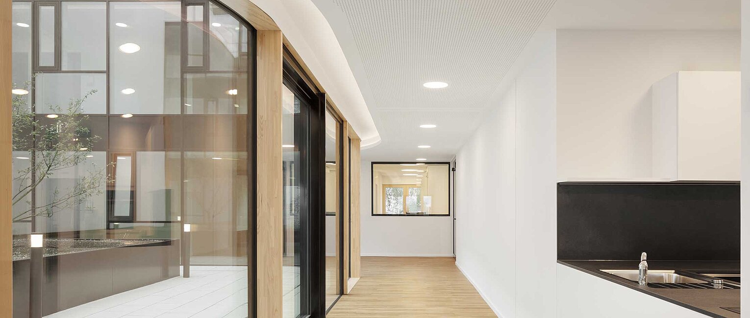 Moderner heller Flur mit Holzboden, weißen Wänden und großen Fenstern, die Blick in einen Innenhof mit Pflanzen bieten.