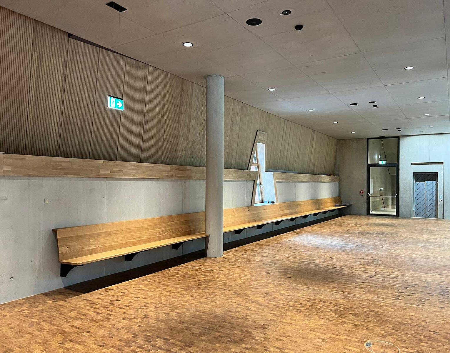 Leerer Flur mit langer, hölzerner Sitzbank, Betonwand und Decke mit Einbaustrahlern.