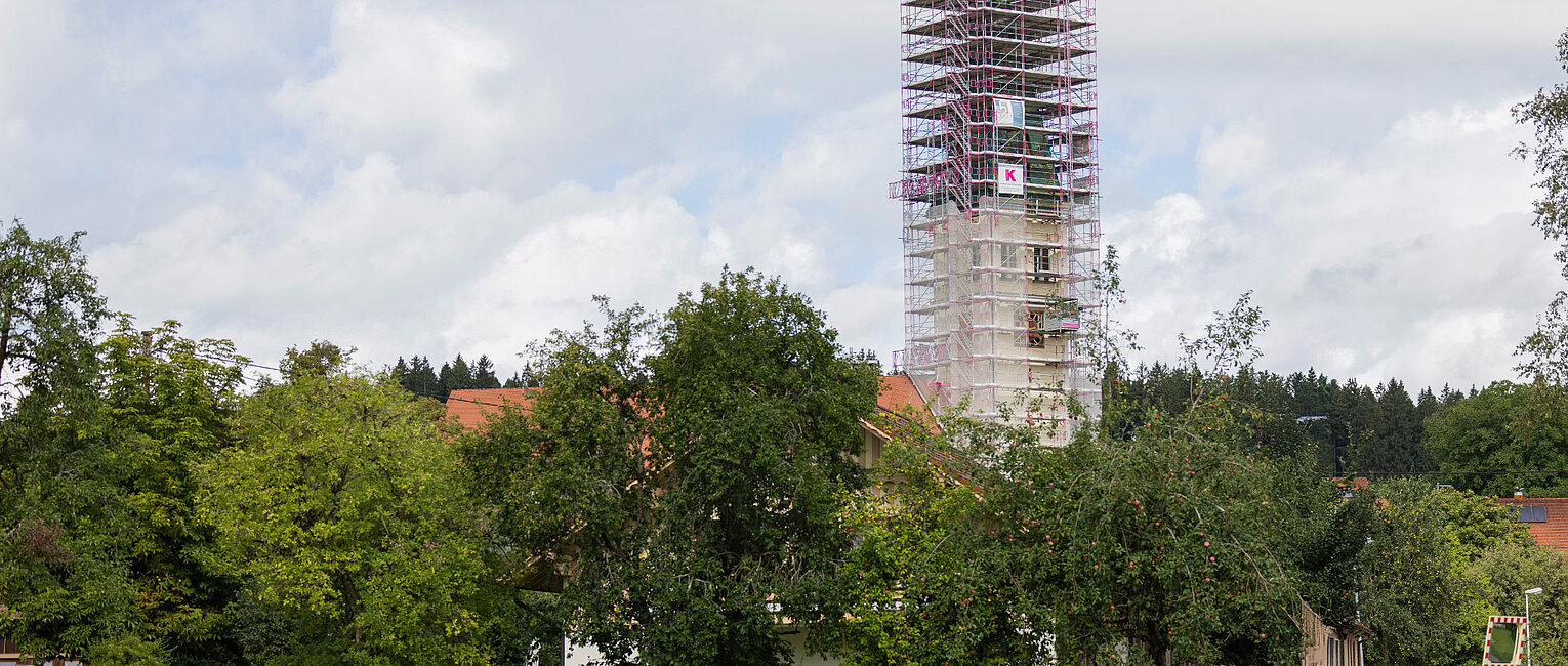Kirchturm im Dorf mit Gerüst und Bauarbeiten, umgeben von Bäumen und Wiesen unter bewölktem Himmel.