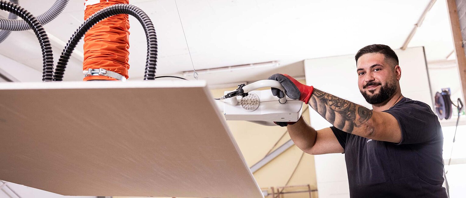 Ein Mann mit Tattoos und Handschuhen bedient in einer Werkstatt eine Hebemaschine, die eine große Platte anhebt.