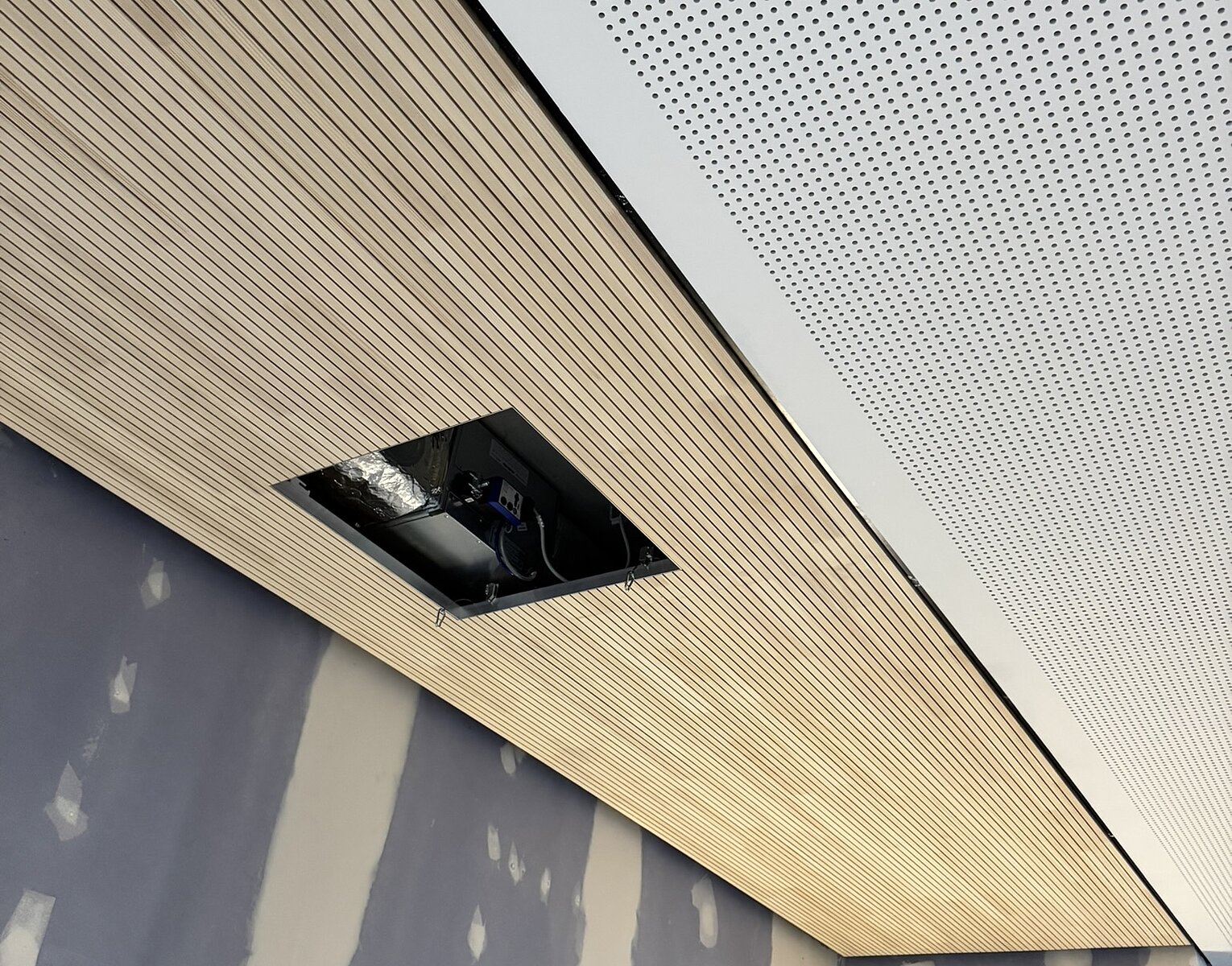 Decke mit zwei verschiedenen Paneelen, eines aus Holzlamellen und das andere perforiert, darunter unfertige Wände mit grau grundiertem Gips und Leiter in einer Baustelle.