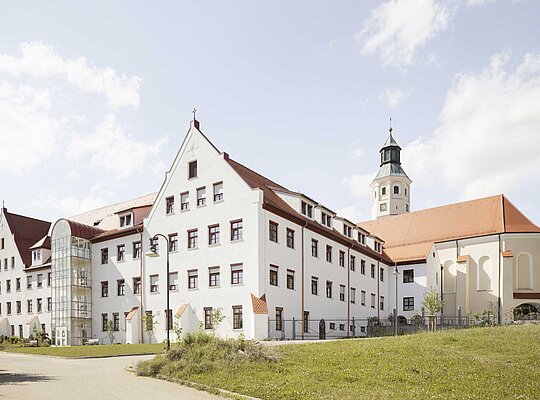 Trockenbau Kaufmann Haus Regenta Großes weißes historisches Gebäude mit rotem Dach und Turm, umgeben von grünem Gras und blauem Himmel mit Wolken.