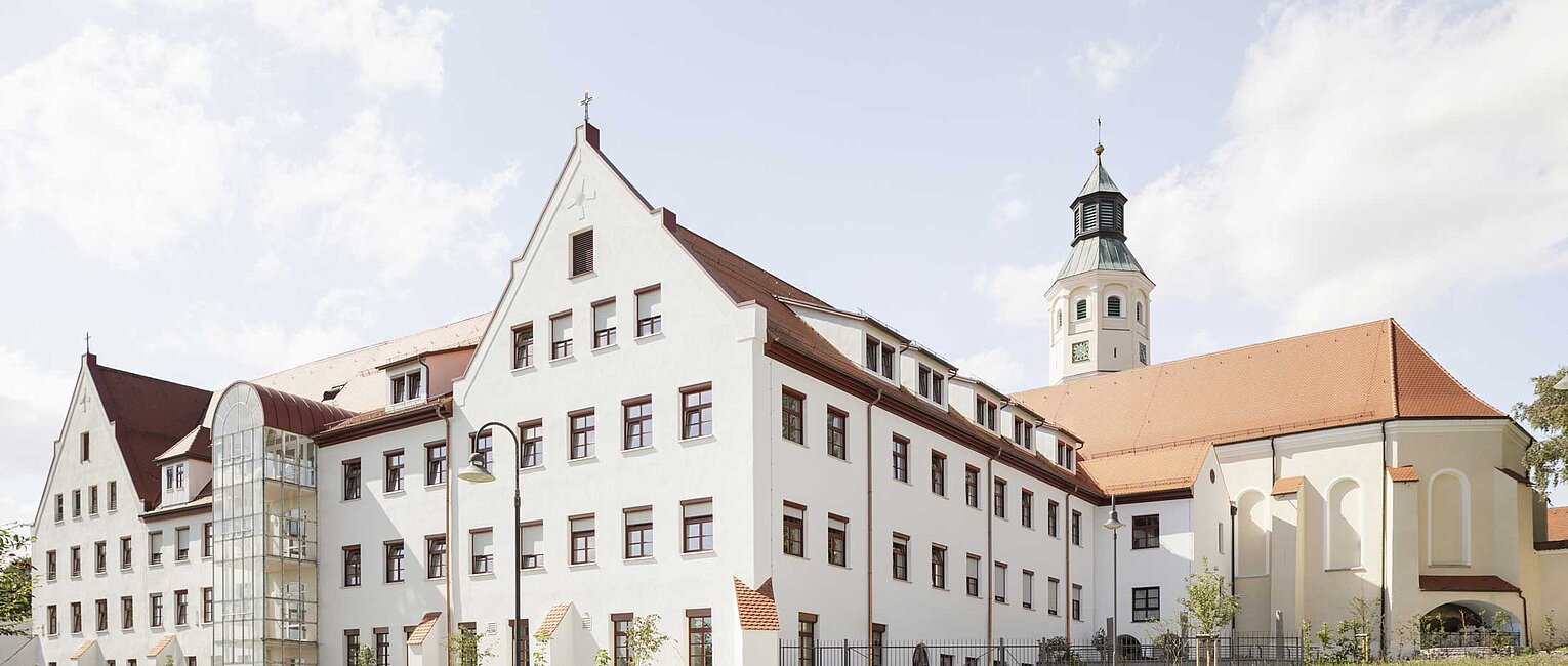 Großes weißes historisches Gebäude mit rotem Dach und Turm, umgeben von grünem Gras und blauem Himmel mit Wolken.