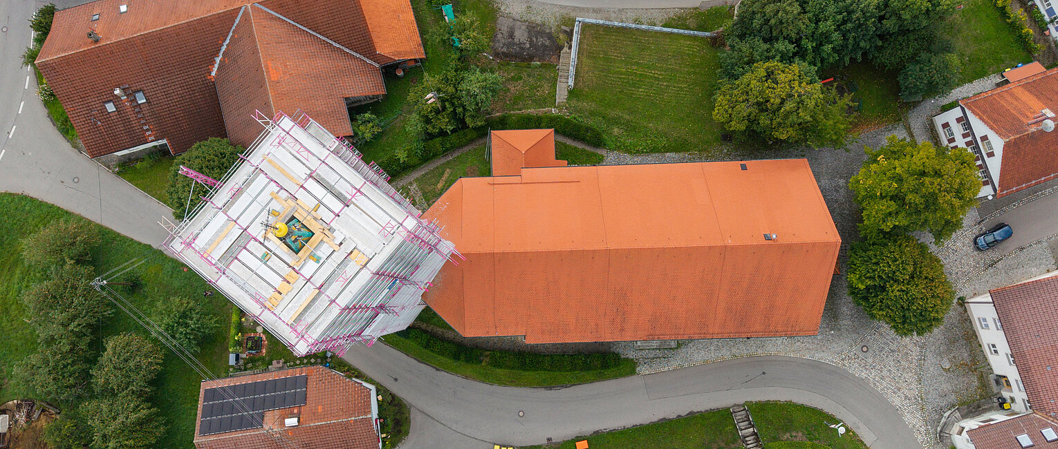 Luftaufnahme eines Gebäudekomplexes mit roten Dächern, einer Straße und grünen Flächen, ein Turmgerüst auf einem der Dächer ist sichtbar.
