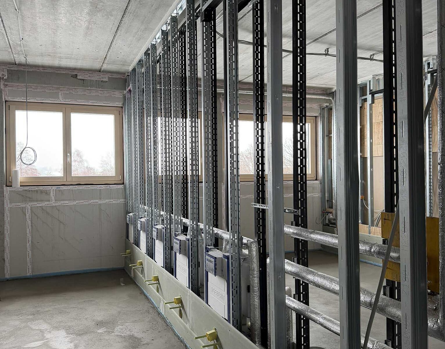 Innenraum einer Baustelle mit unvollständigen Metallständerwänden und sichtbarem Betonboden unter einer Betondecke, Fenster an einer Wand lassen Tageslicht herein.