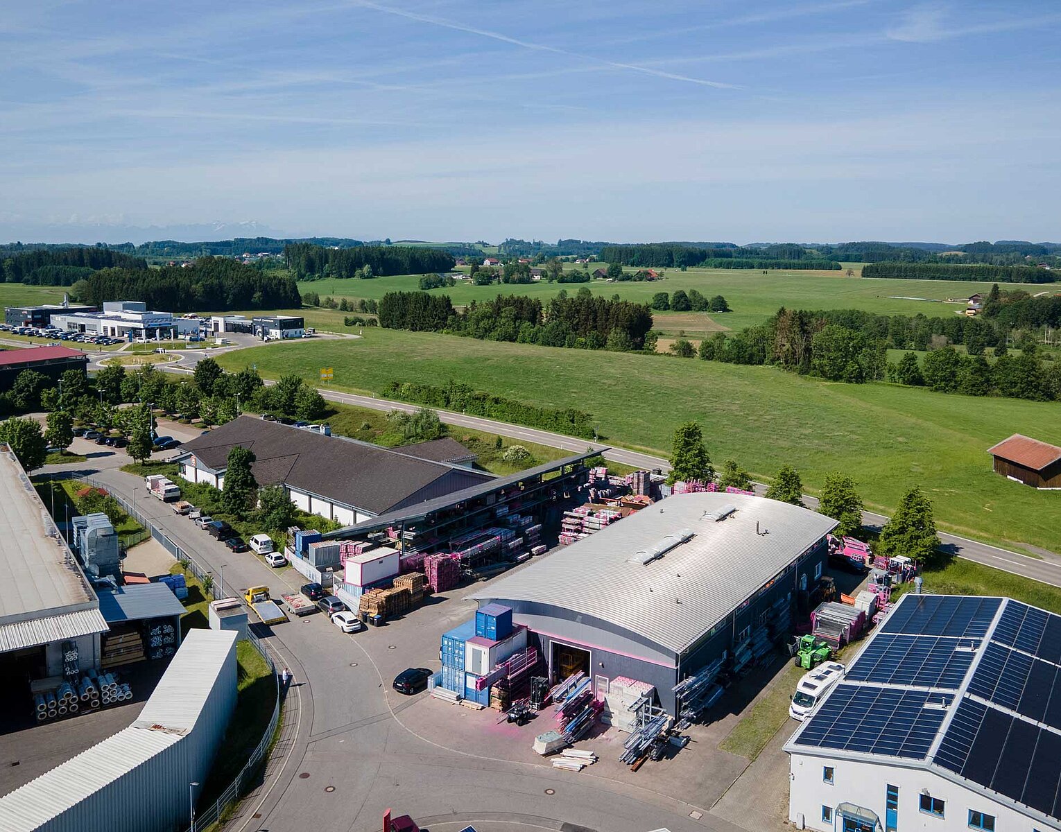 Gerüstbau Kaufmann Luftaufnahme eines Industriegebiets mit mehreren Gebäuden, Parkplätzen, Lastwagen und grünen Feldern im Hintergrund unter blauem Himmel.