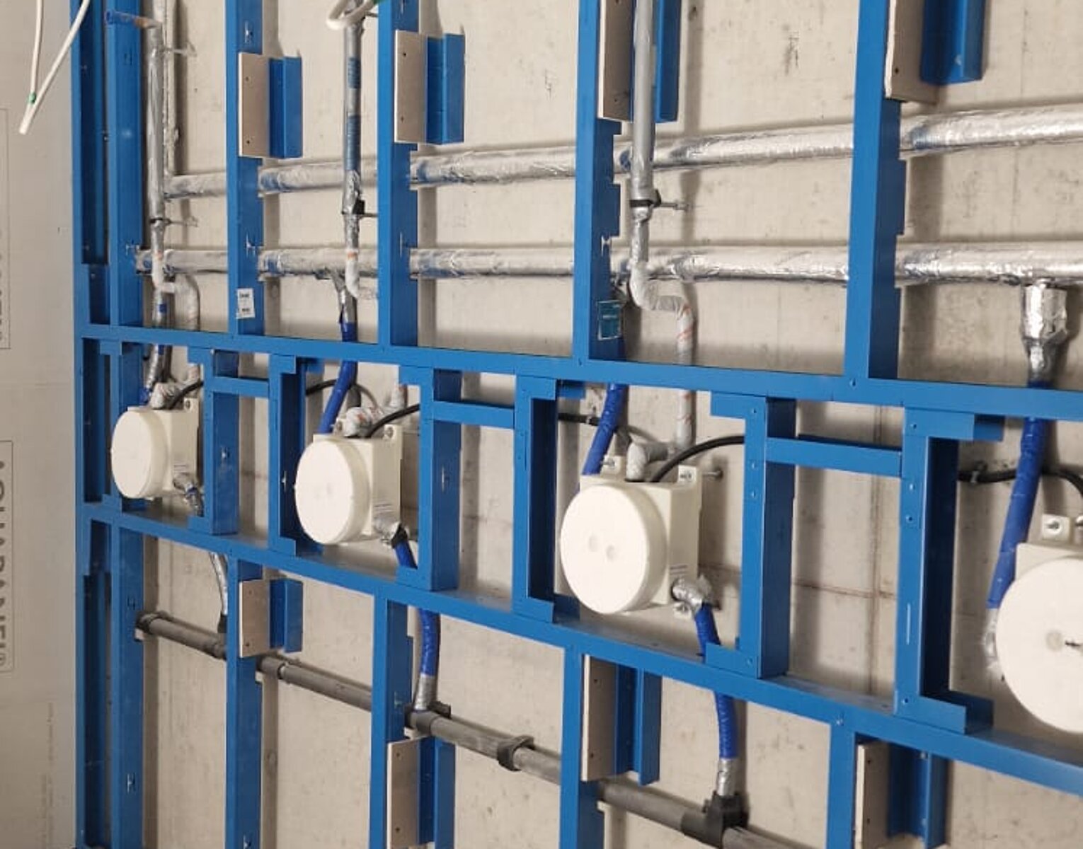 Blaue Metallrahmen mit befestigten weißen Rohren und elektrischen Komponenten an einer unbehandelten Betonwand in einem Rohbau.
