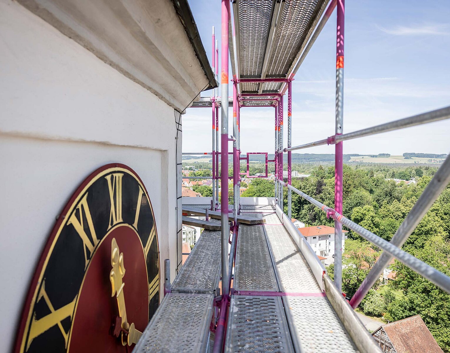 Große Ziffernblattuhr an einem Turm mit Gerüst und Landschaft im Hintergrund bei klarem Himmel.