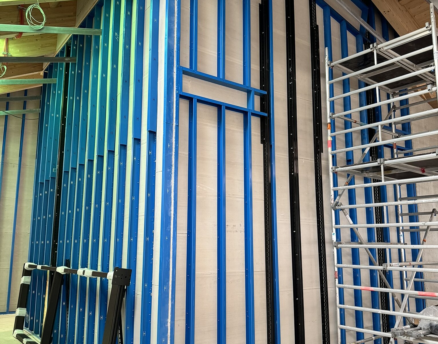 Innenaufnahme einer Baustelle mit blauen Metallständern für Wandverkleidung und einem eingerüsteten Bereich unter einem schrägen Holzdach.